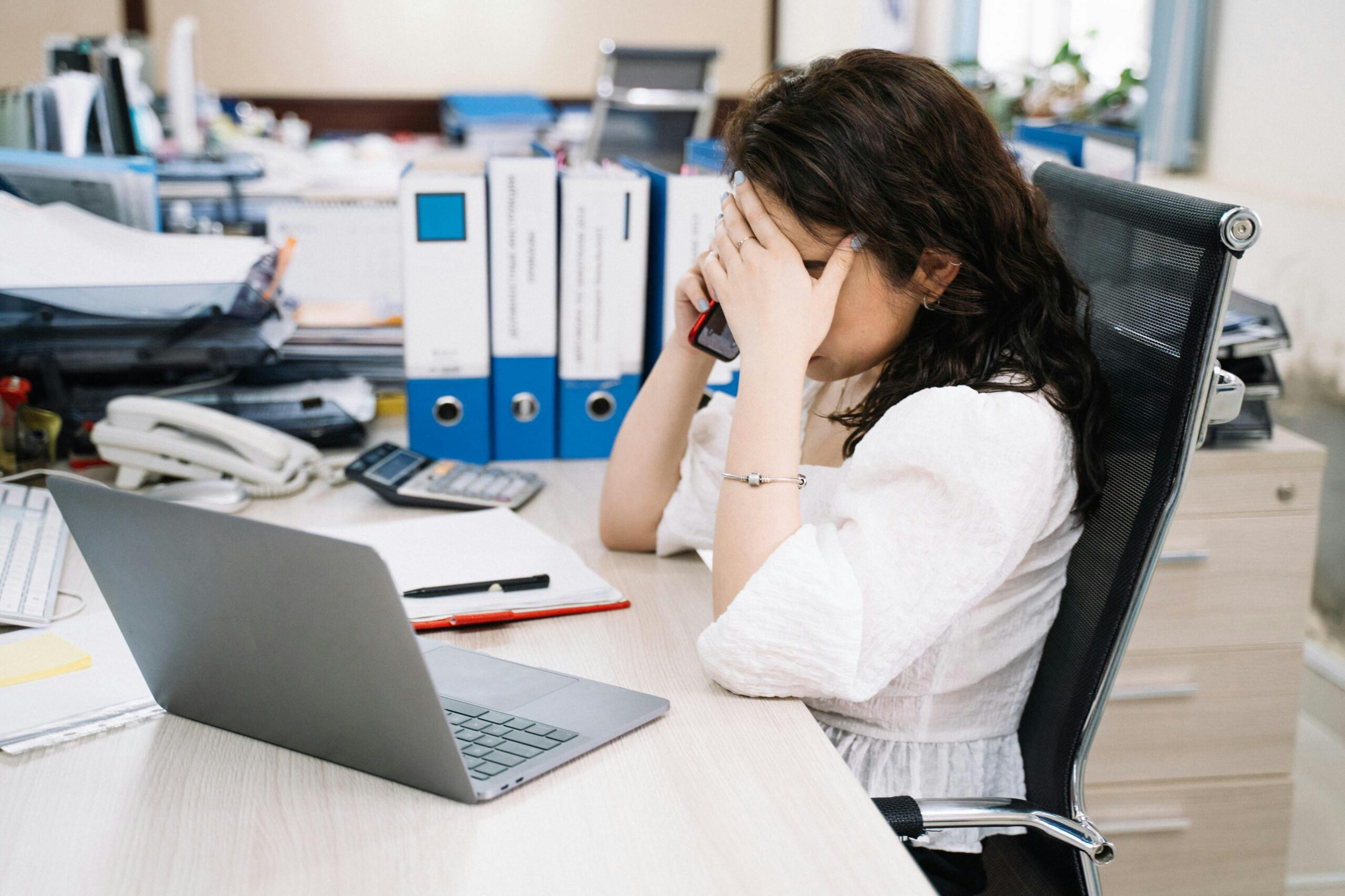 A woman in an office sitting at a desk, visibly stressed, using a phone. Workplace stress concept.